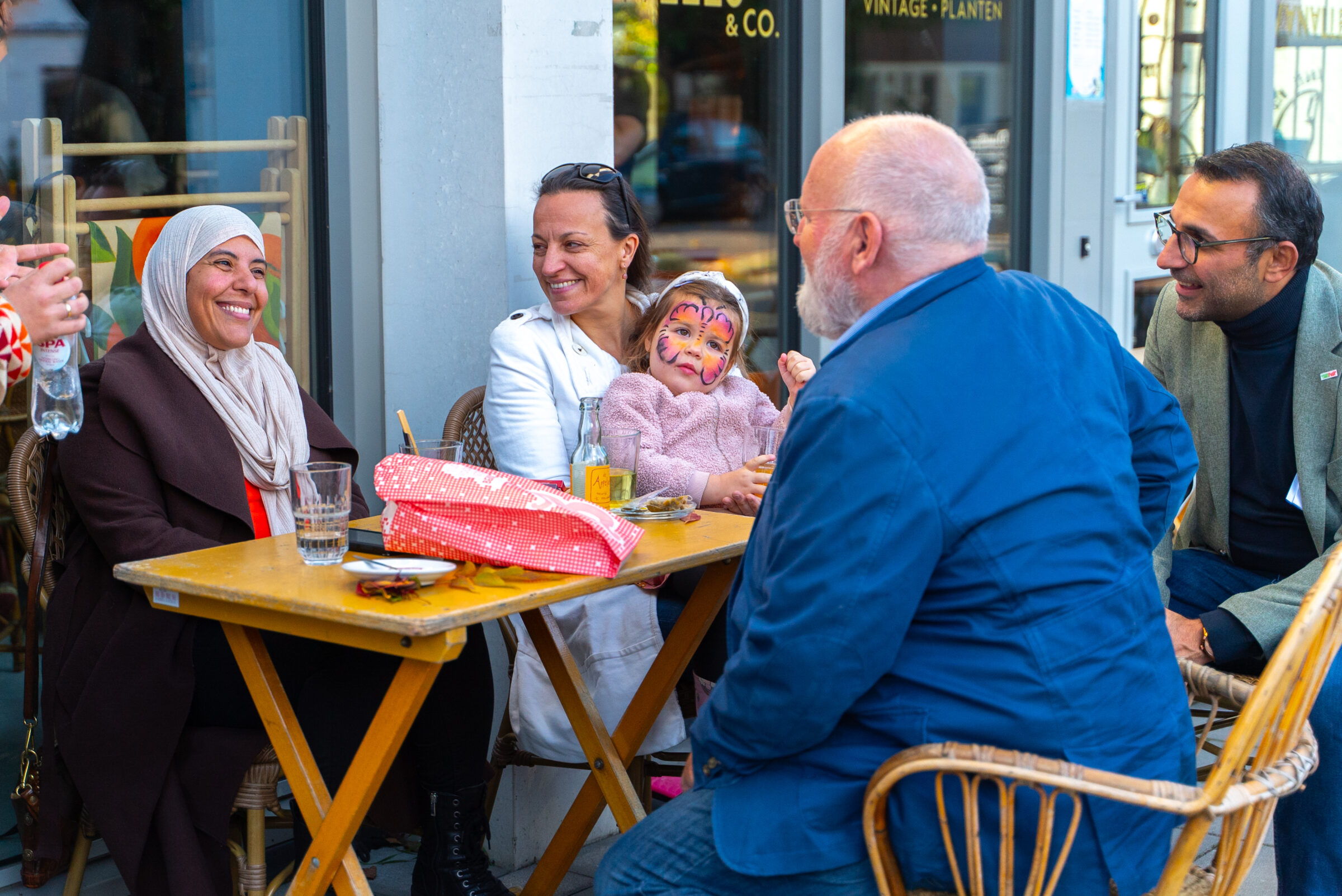 Frans Timmermans zit buiten aan een tafel en praat met twee vrouwen en een man. Een van de vrouwen heeft een kind in haar schoot met kleurige schmink op haar gezicht. 