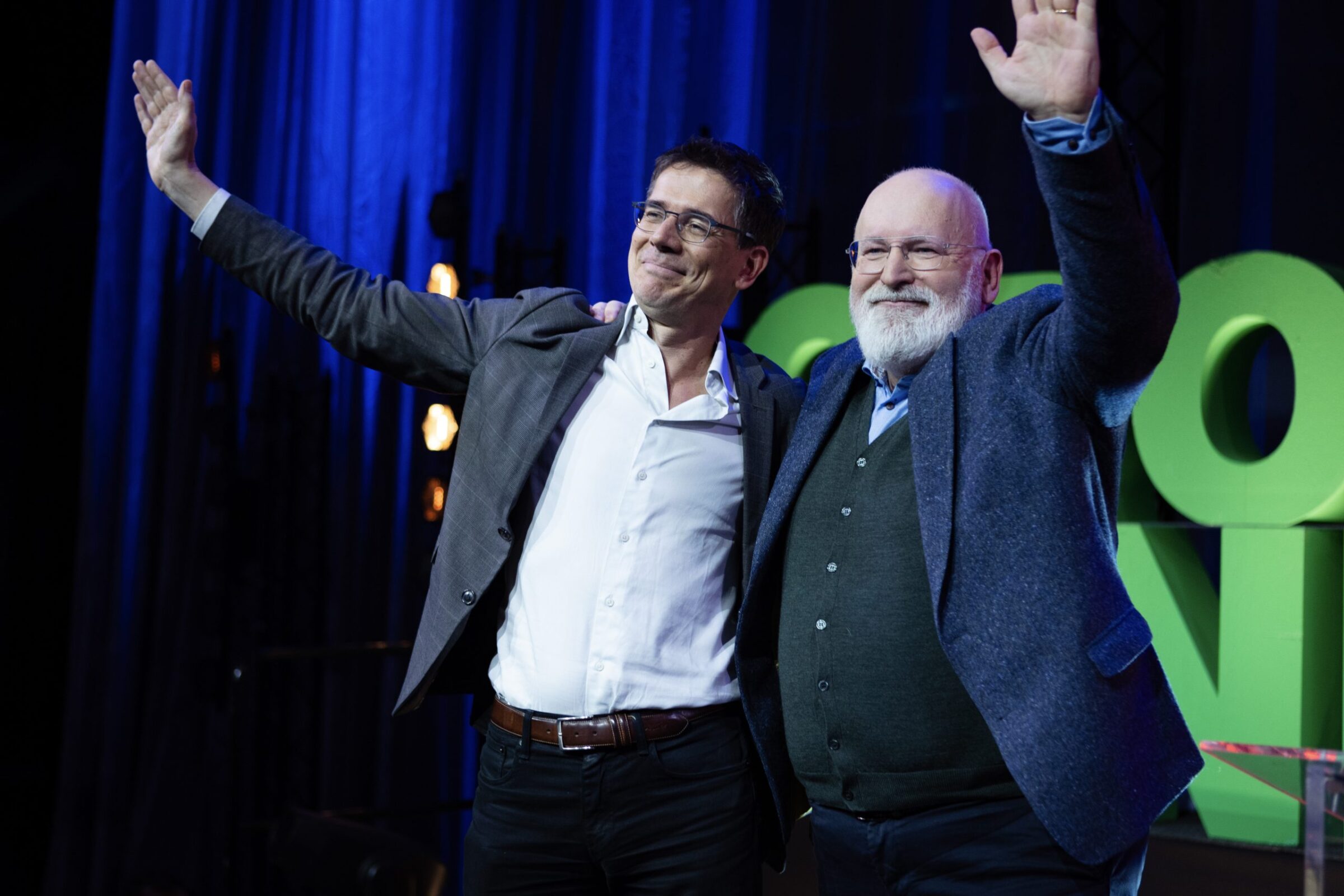 Bas Eickhout en Frans Timmermans op het podium van het GroenLInks-PvdA congres. Ze hebben hun handen om elkaars schouders en in de lucht.